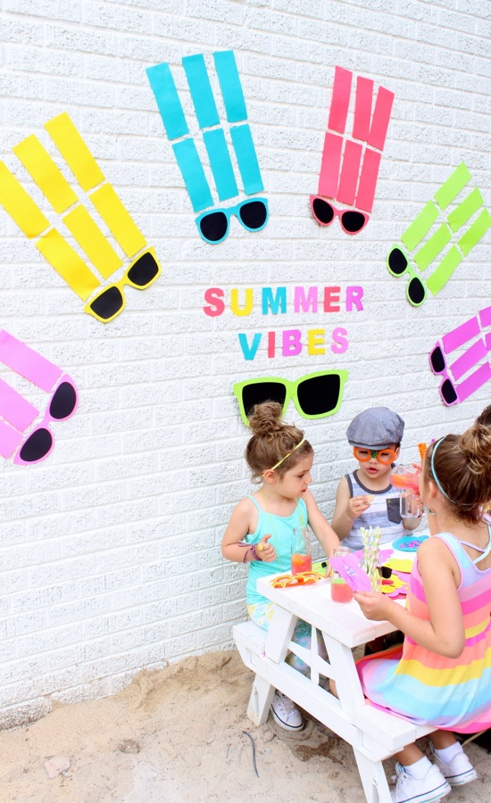 summer-vibes-paper-backdrop-colorful-sunnglasses-arched-like-a-rainbow-kids-playing-and-crafting-on-a-white-picnic-bench